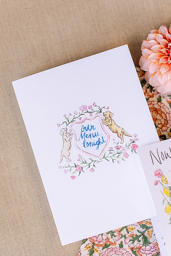 Wedding menu card with watercolor floral and dog illustrations on patterned fabric, set on a linen tablecloth with a blush dahlia nearby