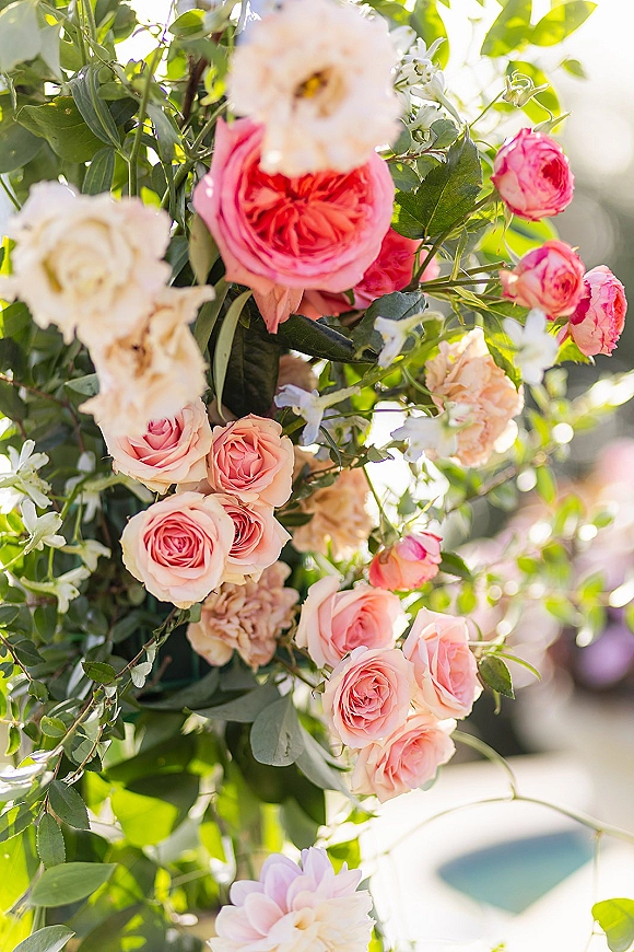 Wedding floral arrangement of pink and peach roses with greenery and a floral vine on a ceremony floral pillar in soft sunlight outdoors
