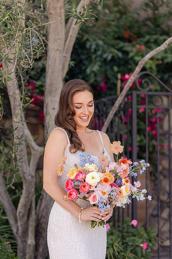 Bridal portrait of a smiling bride holding bouquet, wearing a sequin spaghetti strap gown by a wrought iron gate in lush garden greenery