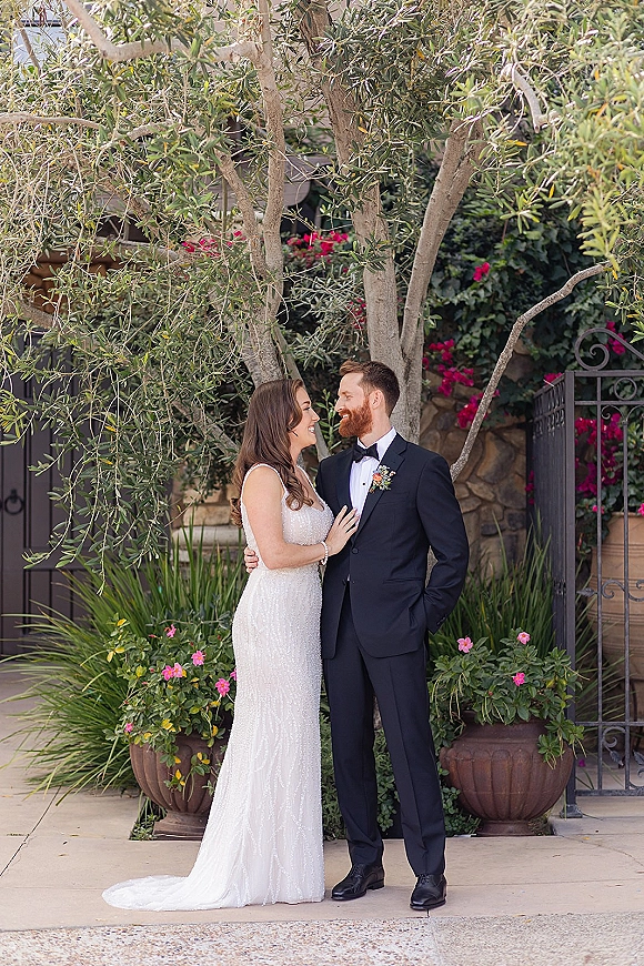 Couple portrait of bride and groom smiling, her hand on his chest, in a garden by an olive tree with stone wall behind