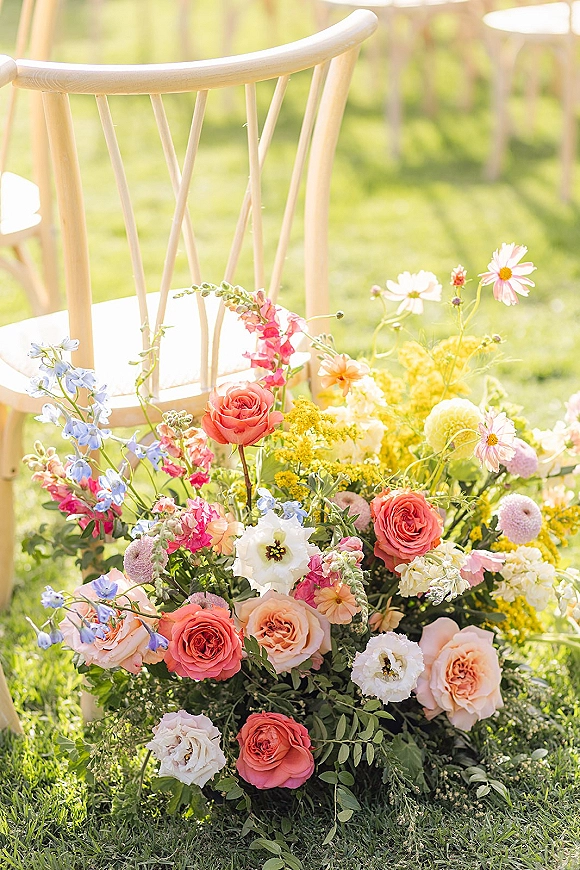 Ceremony aisle flowers in a ground floral arrangement beside a wooden chair, with roses, delphinium and greenery on a sunlit lawn