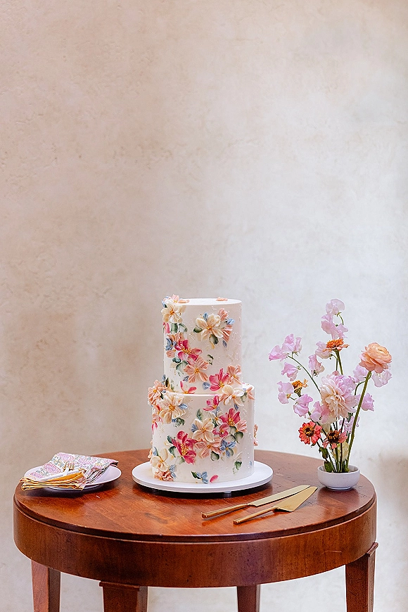 Wedding cake with two tier wedding cake design, hand-painted floral buttercream on a stand with gold knife, server, plates on a round wooden table