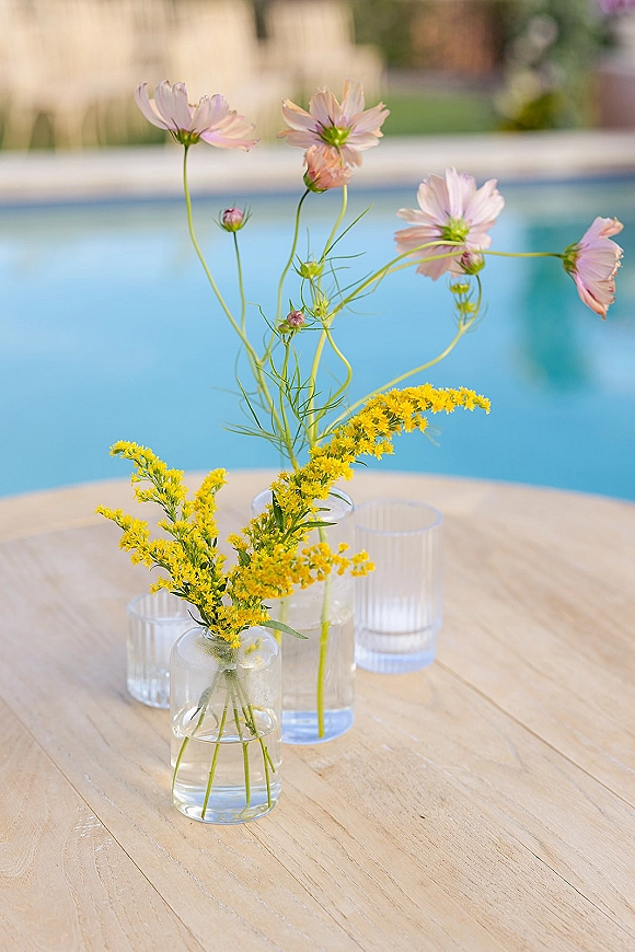 Wedding centerpiece with wildflower wedding centerpiece stems in clear glass bud vases and ribbed votives on a wood poolside table with lawn chairs