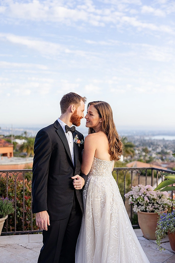 Couple portrait of bride and groom embracing on a terrace patio, her strapless lace dress beside his tux, with city skyline and ocean view behind