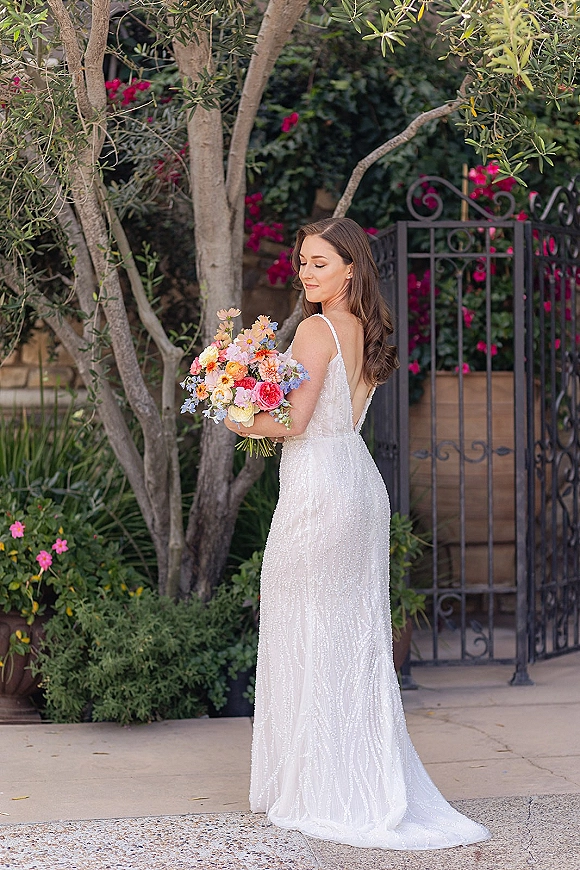 Bridal portrait of a bride holding bouquet in a beaded low-back gown, looking over her shoulder on a garden walkway by a wrought iron gate