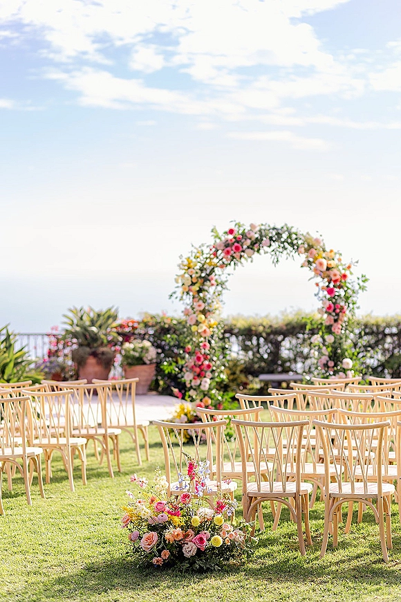 Ceremony setup with an outdoor wedding ceremony floral arch and grounded blooms, wooden chairs on a lawn facing an ocean view under clouds