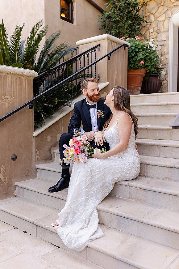 Couple portrait of bride and groom sitting on steps, she holds a colorful bouquet and looks at him beside greenery and railing.