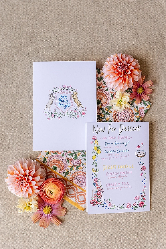 Invitation suite with wedding menu card, floral-illustrated stationery, and pink dahlias with patterned napkins on neutral linen backdrop