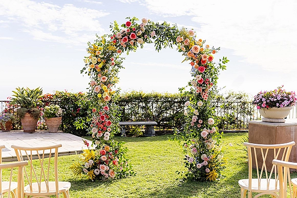 Floral wedding arch with a circular wedding arch frame draped in roses and greenery, set on a garden terrace with wooden chairs and potted plants