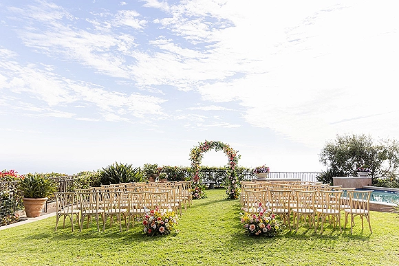 Ceremony setup for outdoor wedding ceremony with a round floral arch, wooden chairs, and aisle flowers facing an ocean-view lawn