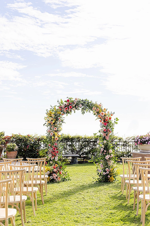 Ceremony setup for outdoor wedding ceremony with a round floral wedding arch of roses and greenery, wooden chairs on a lawn with ocean view