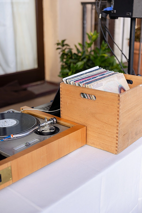 Wedding DJ setup with turntable and vinyl record, wooden crate of sleeves and laptop on a white tablecloth on an outdoor patio