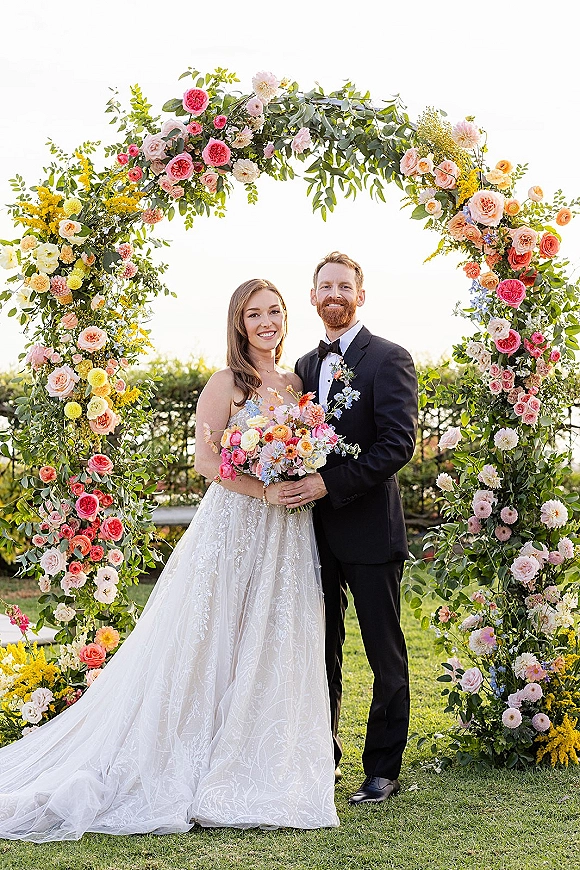 Couple portrait of bride and groom under a circular floral arch, holding a colorful bouquet on a lawn with hedges and open sky