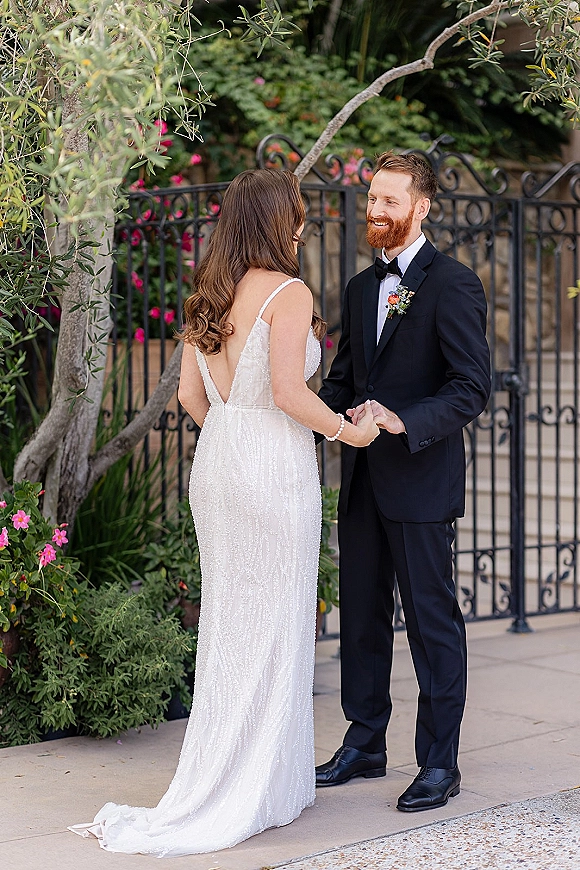 Couple portrait of bride and groom holding hands, smiling by a wrought iron gate in lush garden greenery, her sparkly dress train flowing