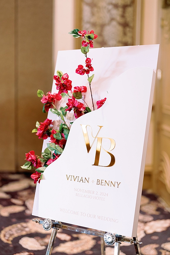 Wedding welcome sign with acrylic design and gold monogram lettering, finished with bright pink florals on an easel in an indoor hallway
