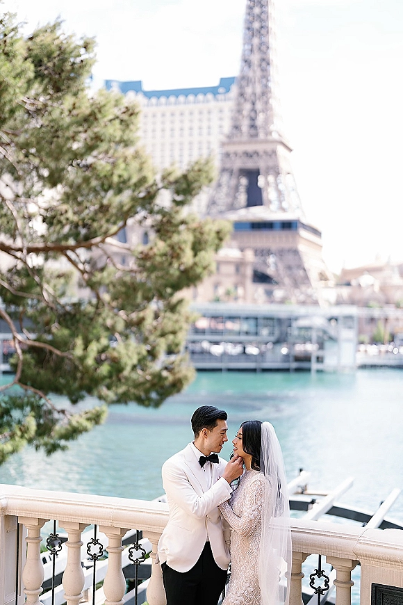 Couple portrait of bride and groom embrace, forehead touching as he cups her face; veil accent on a waterfront balcony with boats and towers