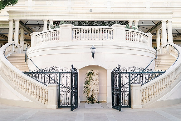 Wedding venue exterior with wrought iron gates and greenery garland framing a white facade, grand staircase, and tiled courtyard entrance
