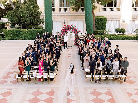 Wedding processional with bride walking down aisle in a cathedral veil and lace train toward a floral arch in an outdoor courtyard ceremony