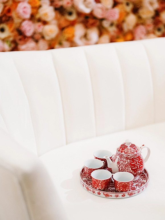 Wedding tea set with red and white porcelain teapot and matching cups on a serving tray, set before a blurred floral wall backdrop