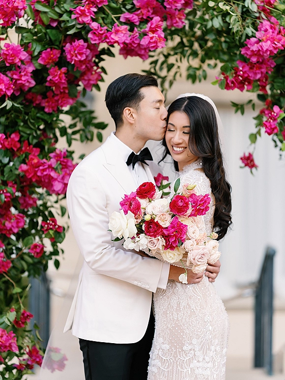 Couple portrait of bride and groom embrace in a forehead kiss, bride holding a bright pink bouquet by a leafy flowering vine wall