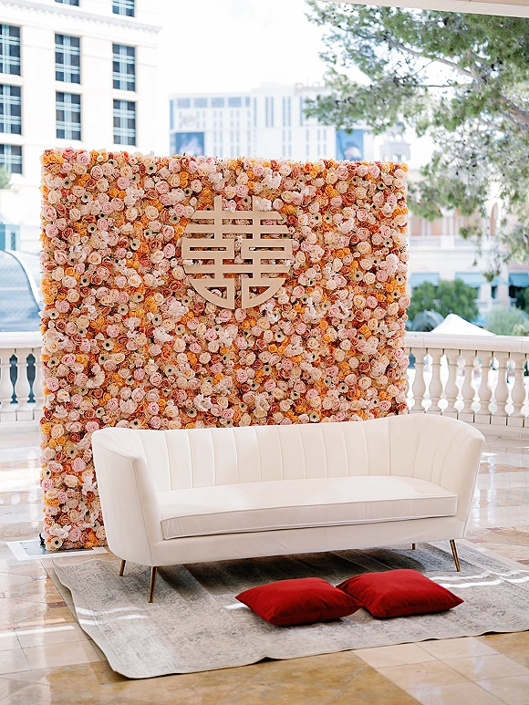 Wedding photo backdrop with a flower wall backdrop of roses and peonies, double happiness sign, white sofa, and red pillows on a terrace