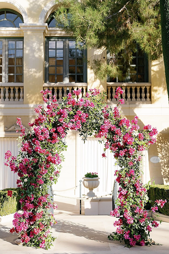 Wedding ceremony arch draped in pink bougainvillea and greenery, set before a villa facade with arched windows and steps