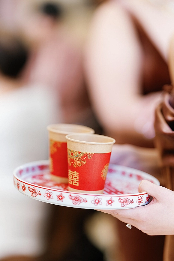 Tea ceremony details with red paper cups on a patterned serving tray, double happiness symbol visible as hands with a ring hold it indoors