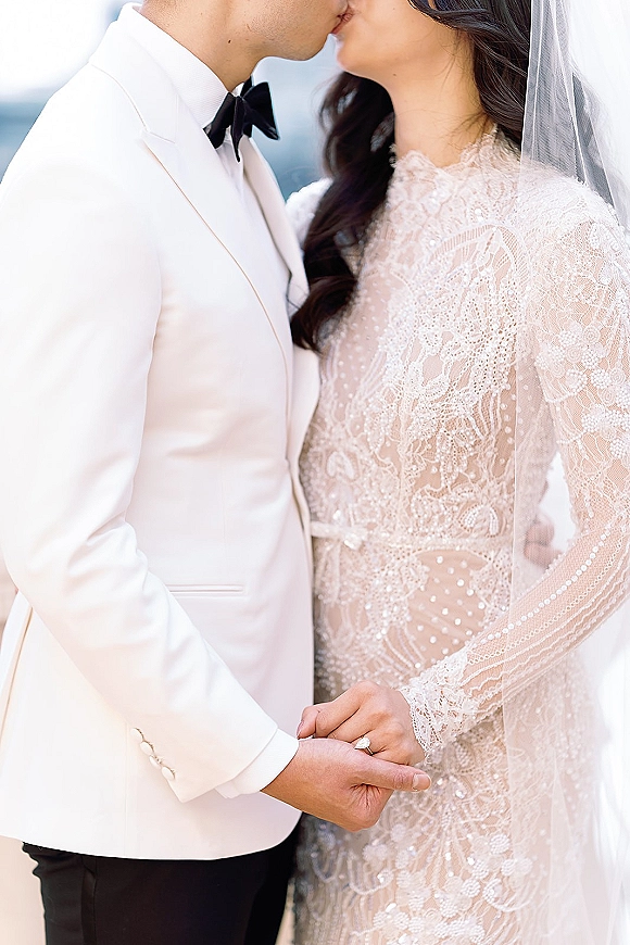 Wedding kiss as bride and groom kissing close up, holding hands with wedding ring visible, lace dress and veil, white tux under open sky