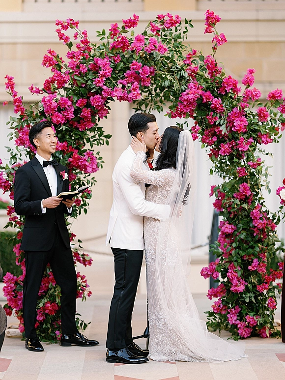 Ceremony kiss as bride and groom kiss under a pink floral arch, her long veil flowing beside him in a white tuxedo jacket by stone columns