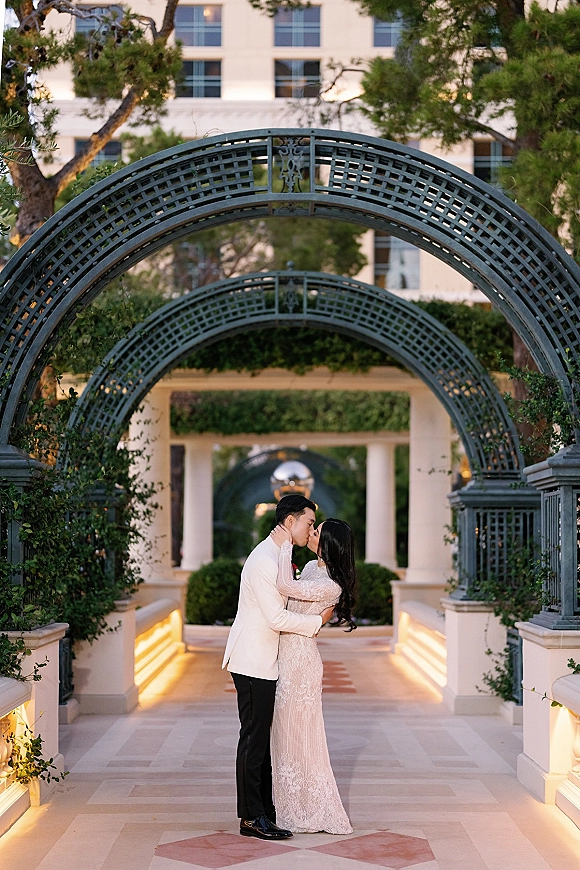 Wedding kiss portrait of bride and groom kissing under a metal garden arbor, lace long-sleeve gown and groom in white dinner jacket at dusk