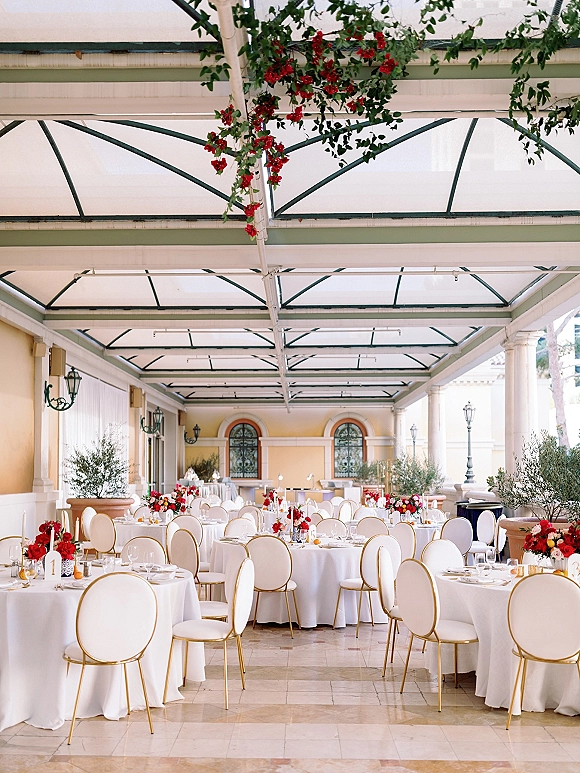 Reception tablescape with round wedding tables dressed in white linens, red rose centerpieces, and taper candles under a glass canopy patio