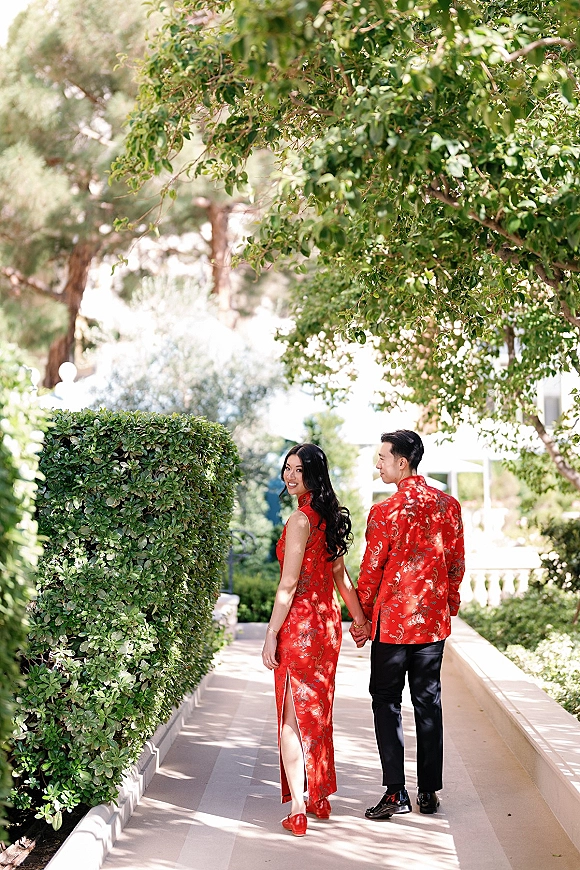 Couple portrait holding hands walking away on a sunlit garden walkway, bride in red cheongsam looking back beside groom in brocade jacket