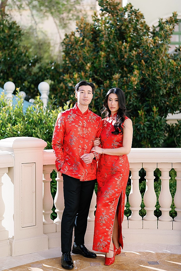Couple portrait in Chinese wedding portrait attire, arm in arm in red embroidered qipao and jacket by a white balustrade in a garden setting