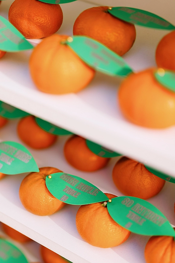 Wedding favor display of orange wedding favors on white shelving, each fruit labeled with leaf-shaped tags and pinned place cards in a neutral setting
