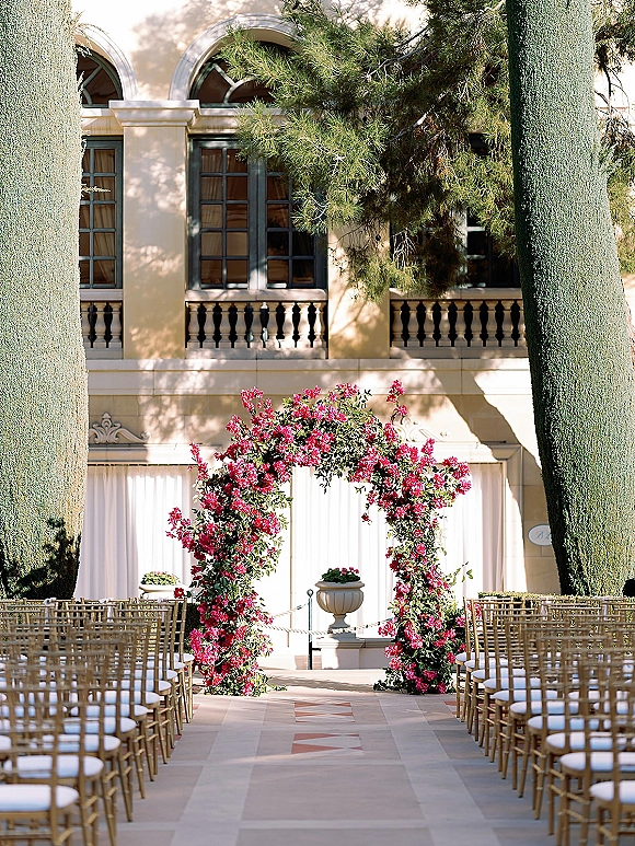 Ceremony setup with floral arch of pink flowers and greenery, gold Chiavari chairs and white runner in a stone villa courtyard backdrop