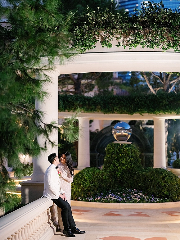Couple portrait of bride and groom posing, bride leaning on groom in formal attire under white-column colonnade with greenery courtyard backdrop