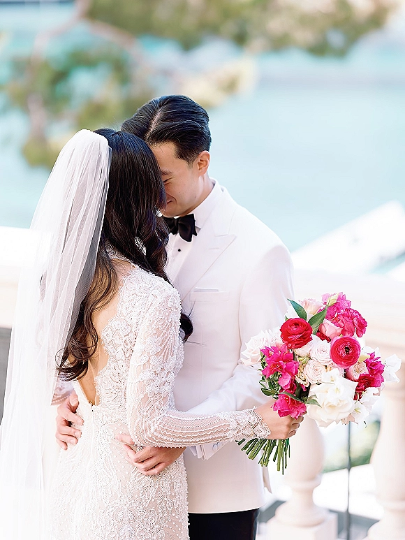 Wedding couple portrait of bride and groom embrace, touching foreheads on an ocean-view terrace with veil, white tux, and pink bouquet