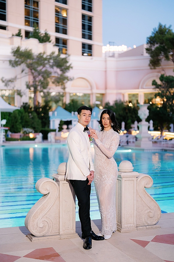 Couple portrait by the pool, bride in a beaded gown holding groom’s lapel as he wears a white tuxedo jacket at twilight resort setting