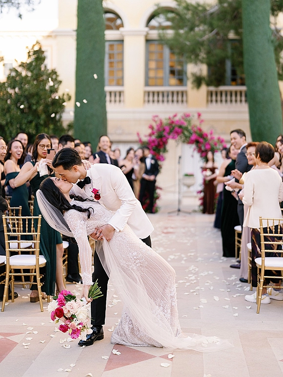 Wedding kiss portrait of bride dipped by groom beneath a fuchsia floral arch, rose petals on the aisle and guests cheering in a courtyard