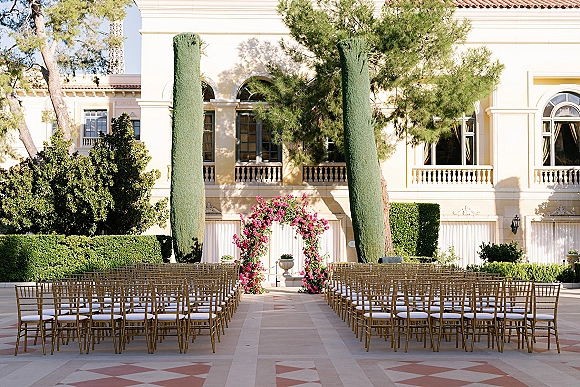 Ceremony setup with a pink floral arch and rows of gold chiavari chairs with white cushions in a villa courtyard with cypress trees