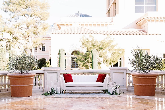 Wedding lounge setup with wedding lounge seating on a white sofa with red pillows, olive trees and pillar candles on a terrace by a stone balustrade