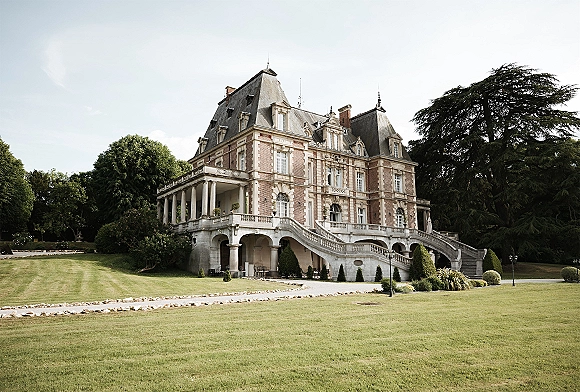 Wedding venue exterior of a chateau wedding venue with a grand staircase and stone balustrade, columned terrace, and manicured lawn entry