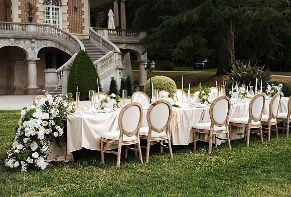 Reception tablescape with a long banquet table, ivory linens, white florals and greenery garland, taper candles, and place settings on a garden lawn near a brick facade