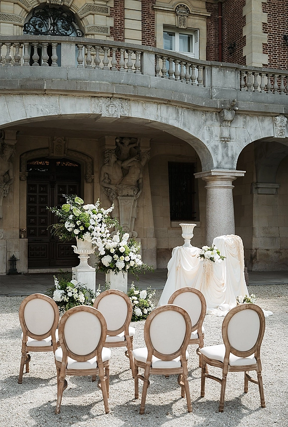Ceremony setup with outdoor ceremony chairs facing a stone archway, framed by white and green urn florals in a gravel courtyard