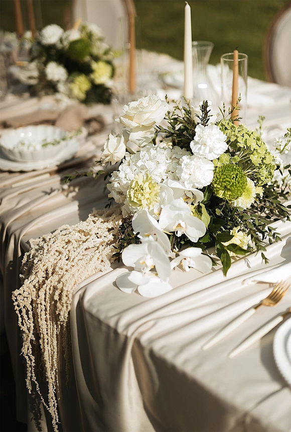 Reception tablescape with wedding table centerpiece of white and green orchids and hydrangea, taper candles, and gold flatware on a sunlit lawn