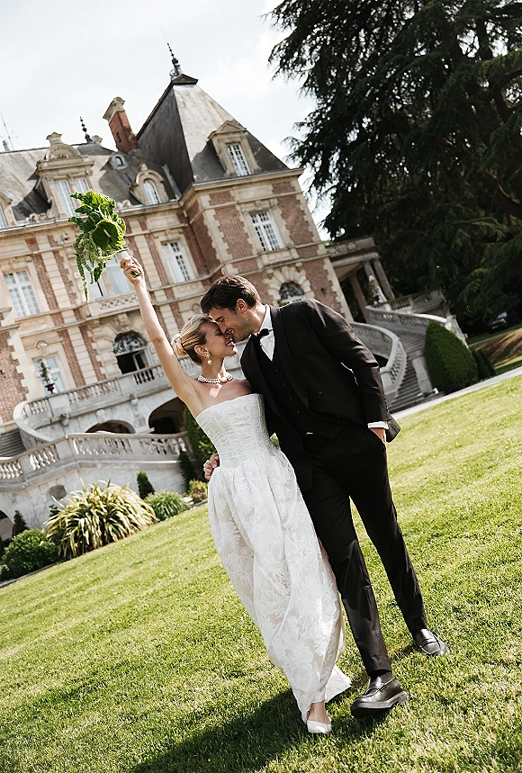 Wedding kiss portrait of bride and groom kissing as she lifts her bouquet, standing on stone steps before a castle backdrop outdoors