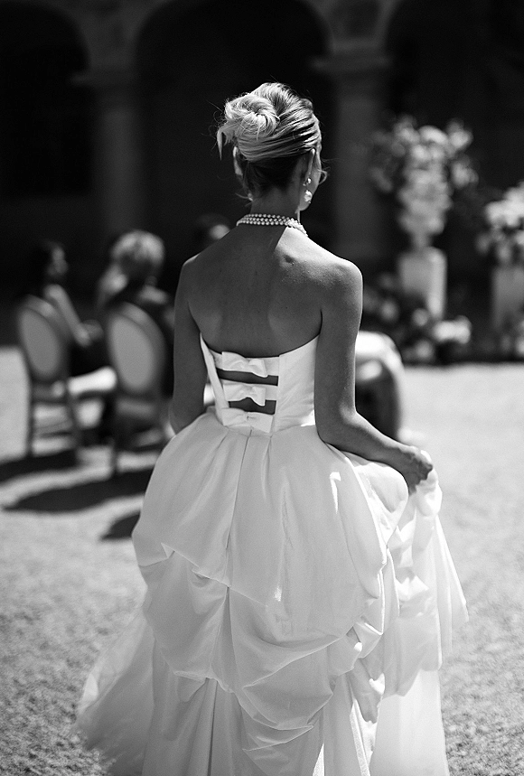 Bridal portrait of a bride from behind in a corset back wedding dress with pearl necklace, walking through a sunlit stone-arch courtyard