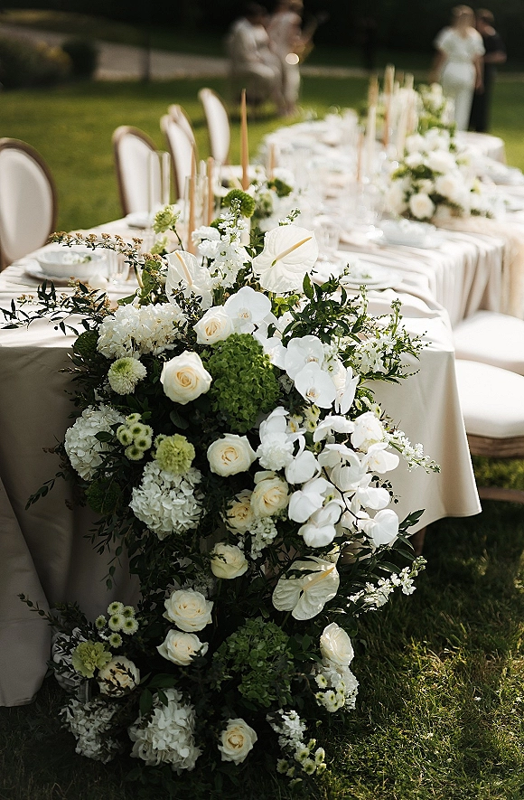 Wedding reception tablescape with head table floral runner of white roses, orchids, hydrangeas and taper candles on an outdoor lawn with guests