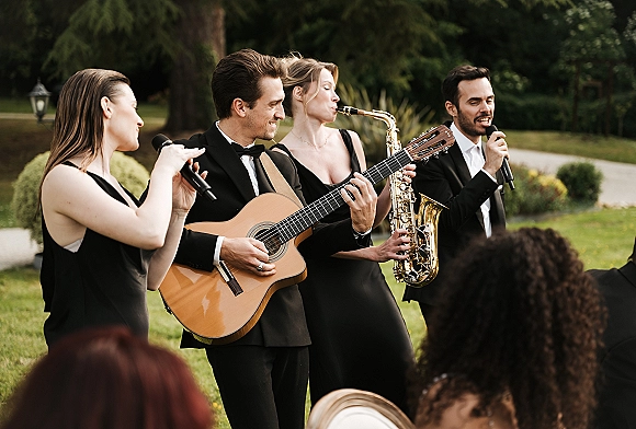 Wedding band performing live wedding music with microphones, acoustic guitar and saxophone, in black tie attire on a garden lawn with guests