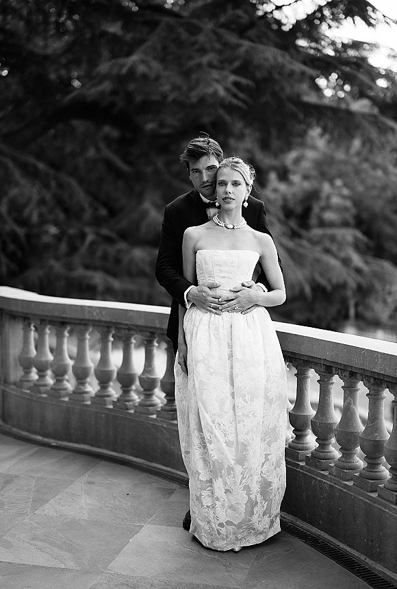 Couple portrait in a black and white wedding portrait as groom hugs bride from behind on a stone balcony, her pearl necklace gleaming